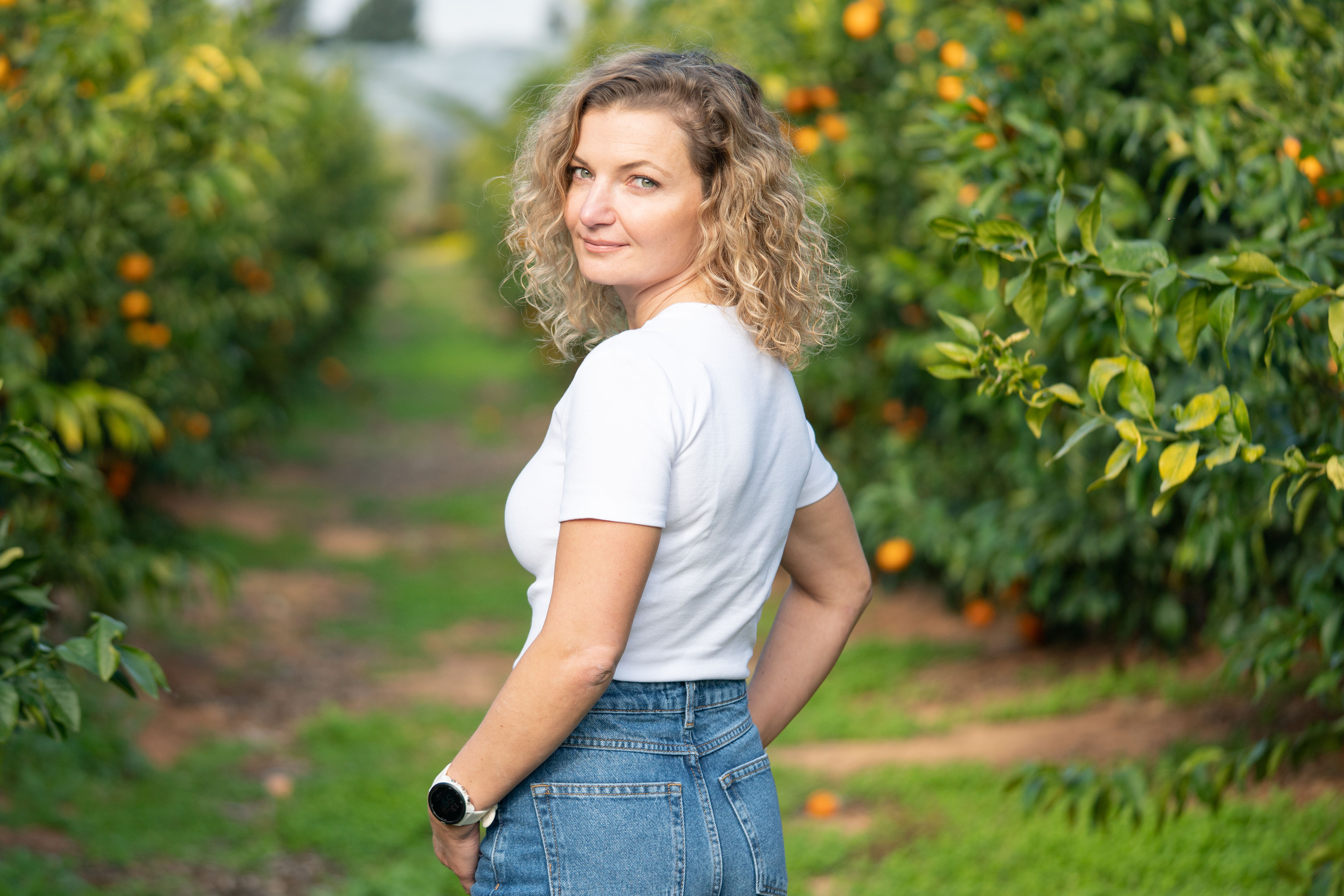 מצולמת עם חולצה לבנה וגינס מסתכלת אחורה על רקע בפרדס