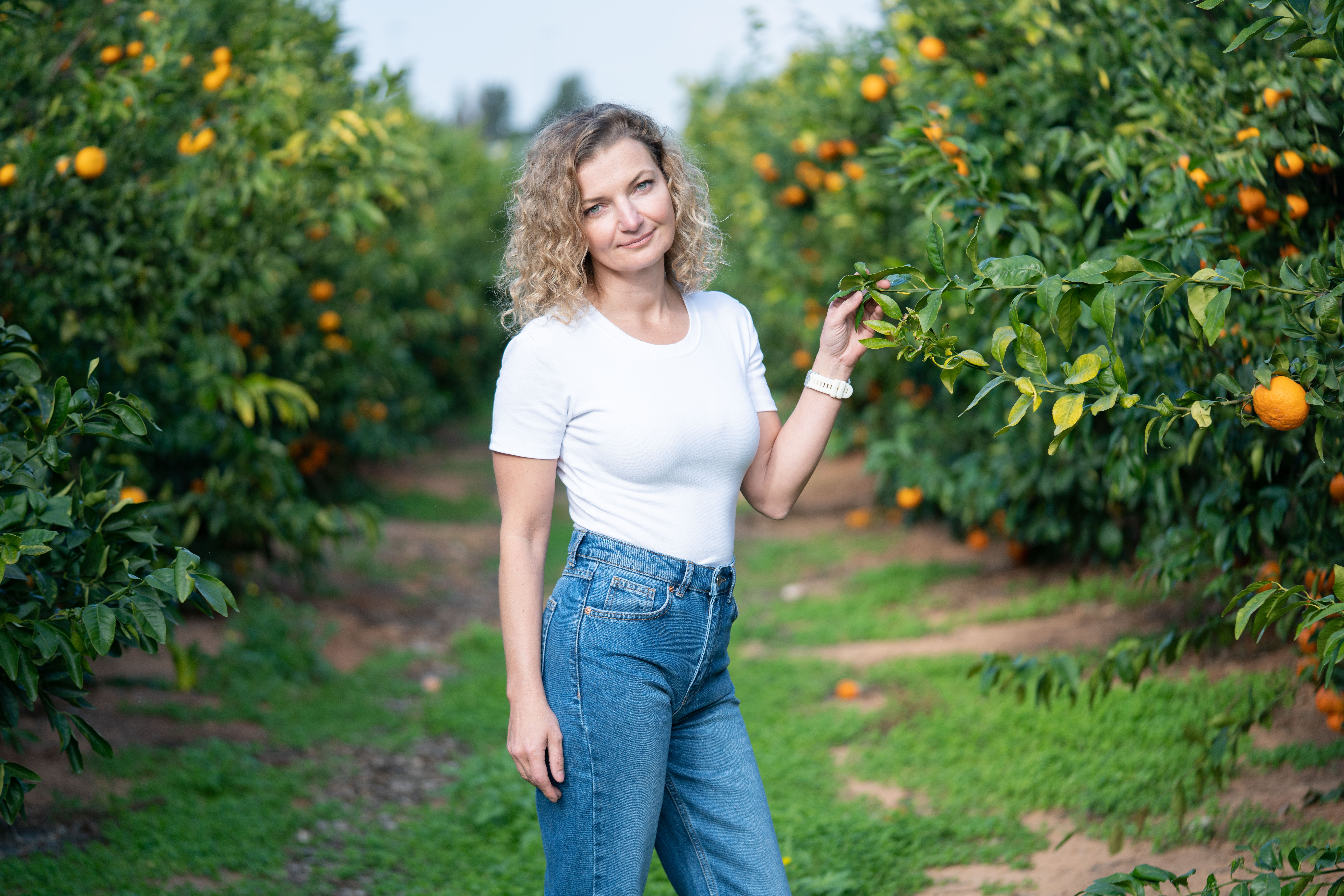 מצולמת עם חולצה לבנה וגינס עומדת ומחזיקה ענף של עץ פרי הדר בפרדס.