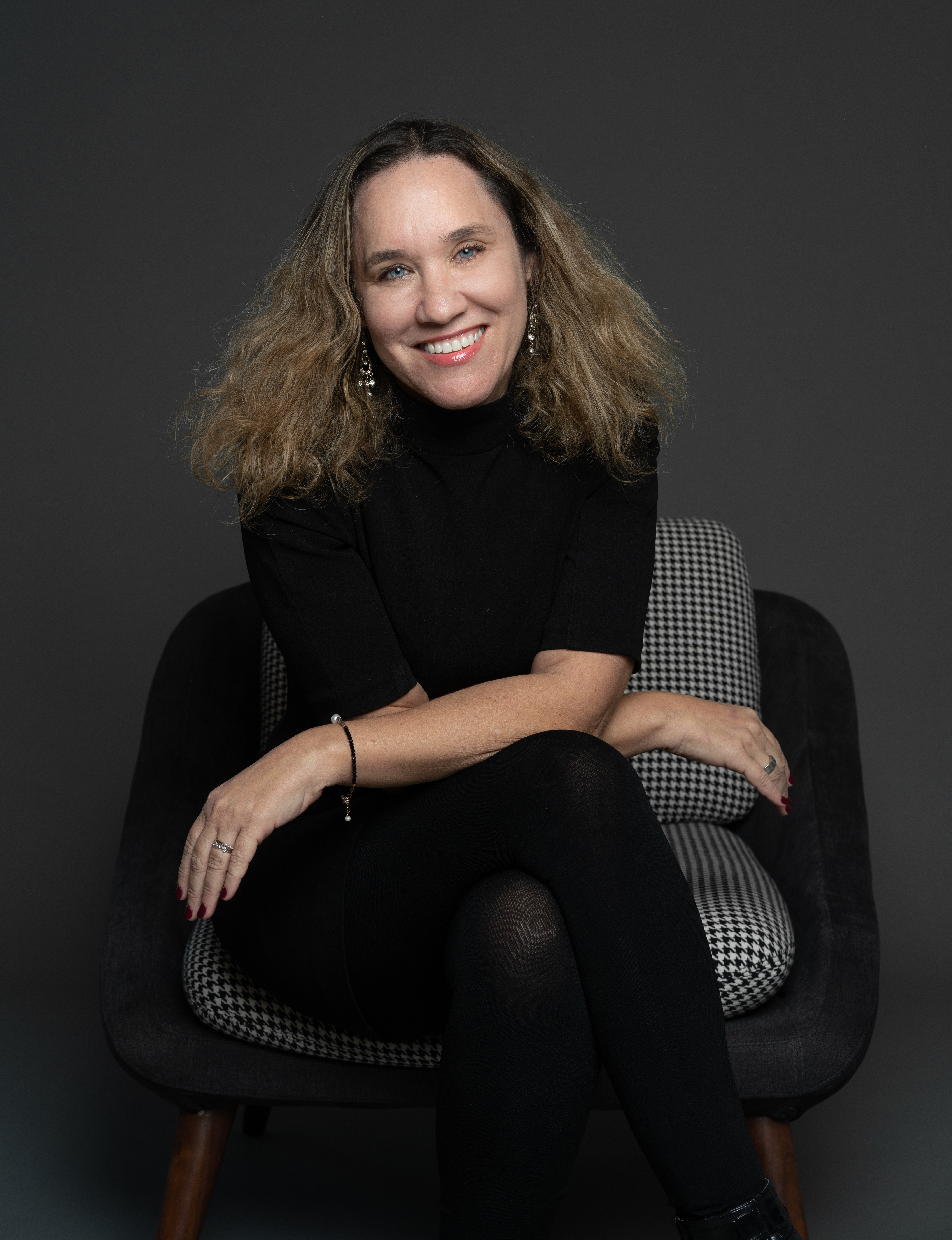 מצולמת בבגדים שחורים יושבת על כורסה אפורה על רקע כהה